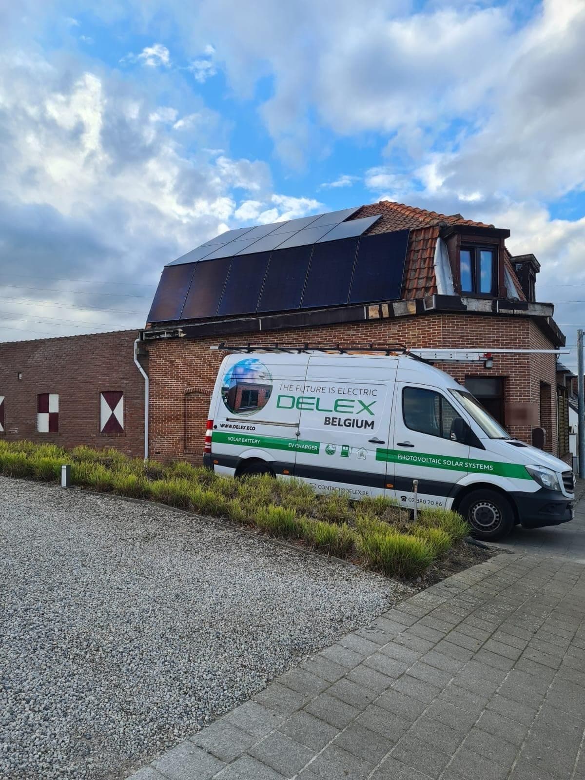 Brick corner house with a mansard roof covered in full-black solar panels, the white DELEX van parked in front